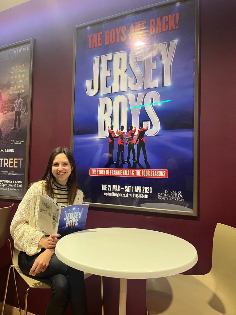 Lauren sitting in front of the advert for Jersey boys holding jersey boys programme advertisement says the boys are back the Jersey Boys the story of Frankie Valli and The Four Seasons Tuesday 21st of March to Saturday first of April 2023 at Royal & Derngate Northampton Telephone 0160 4624 811