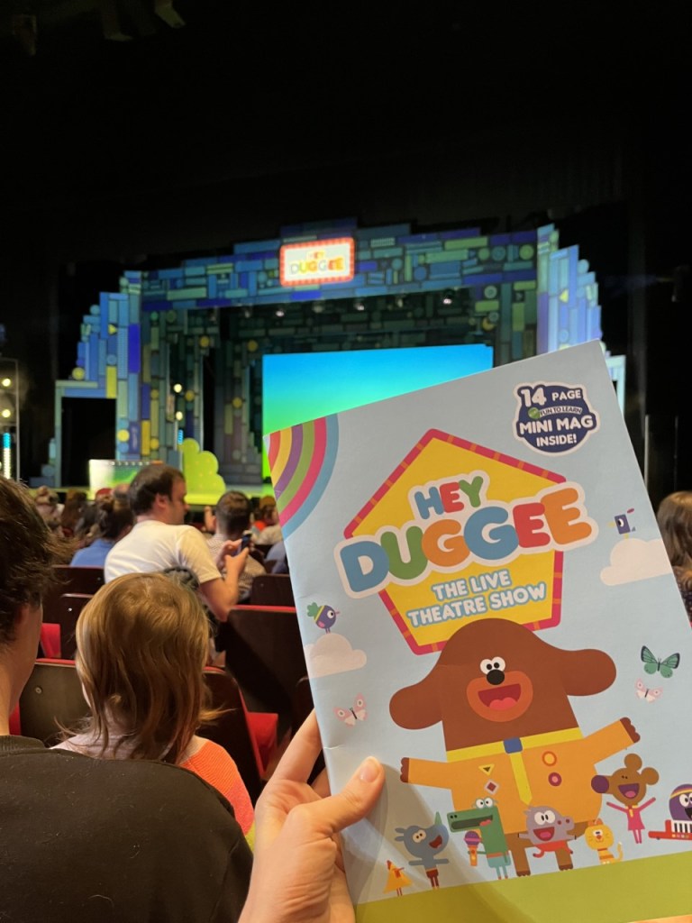 holding the hey duggee programme in front of the bright stage clubhouse